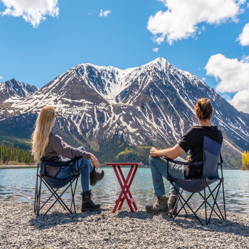 Male and female couple sitting by a lake in a panoramic setting with a mountain view, scene in Kluane National Park with massive snow-capped peaks in the distance.