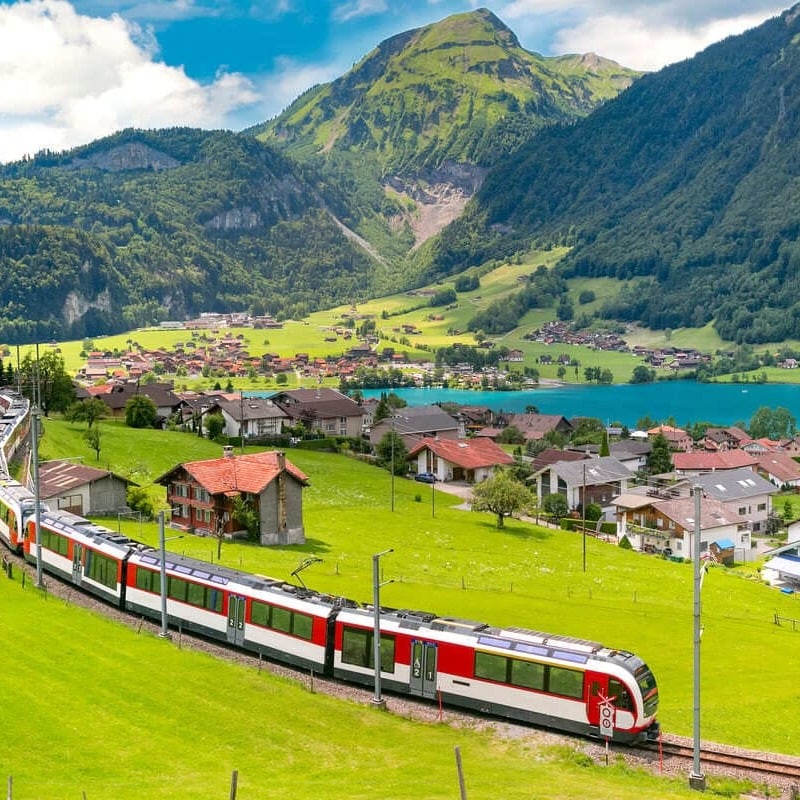 Traditional red tourist train traveling through a picturesque region of Switzerland, next to the Alps, where there is a small Swiss village and an alpine lake, Central Europe
