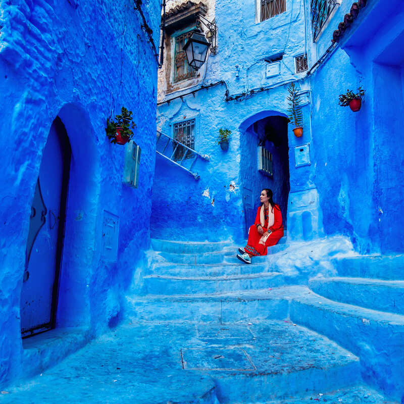 Young Woman Wearing A Red Dress Contrasting Against The Striking Blue Of The Houses In Chefchaouen, The Blue City Of Morocco, North Africa