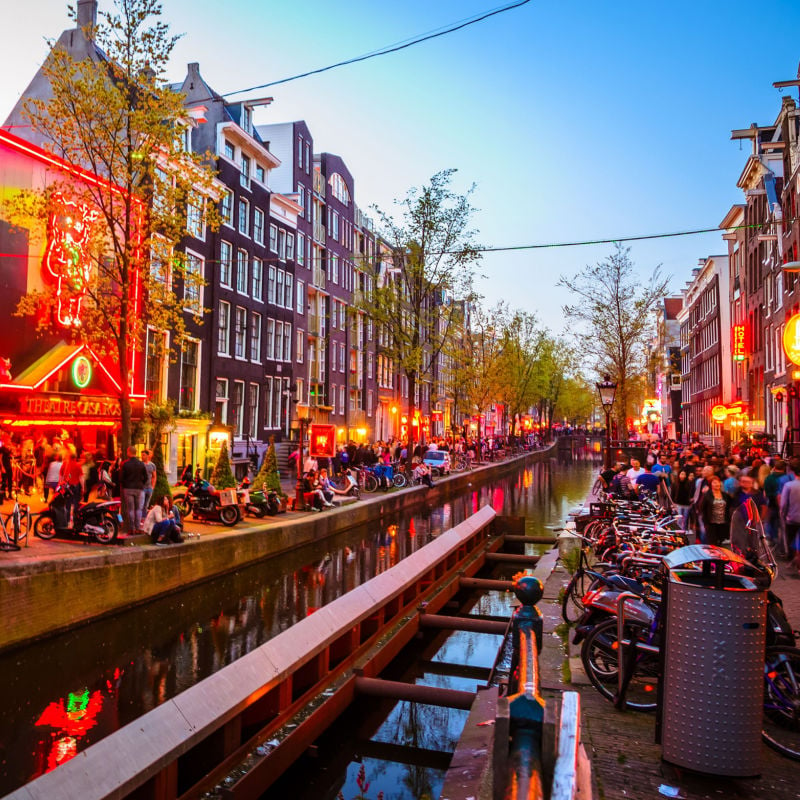 Tourists sit along a canal in Amsterdam with red lights glowing along the buildings as evening approaches